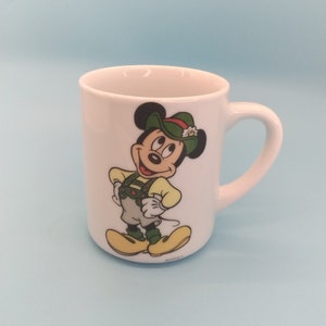 May include: White ceramic mug featuring a cartoon image of Mickey Mouse wearing a green hat and traditional German clothing. The mug has a handle and the text "&copy;Disney" on the bottom.