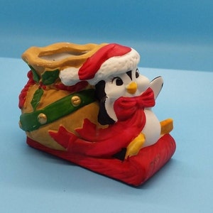 May include: A ceramic Christmas decoration featuring a penguin wearing a Santa hat and a red bow. The penguin is seated on a red sled, with a yellow sack behind it. The sack is decorated with green and gold accents.