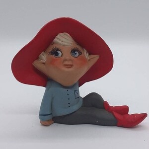 May include: A ceramic figurine of a sitting elf wearing a red hat and blue shirt. The elf has blonde hair, blue eyes, and rosy cheeks.