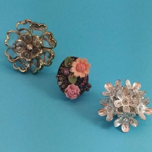 May include: Three floral hair clips in different styles. The first clip is gold with a clear flower design. The second clip is round with a dark background and colourful flowers. The third clip is silver with a cluster of white flowers.