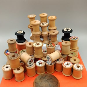 May include: A selection of vintage wooden thread spools in various sizes and shapes. Some spools have colourful labels, while others are plain. A dark brown, rounded object sits at the bottom. The spools are arranged on an orange surface.
