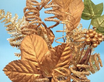 May include: A gold-colored floral arrangement with leaves, sprigs, and curly stems. The arrangement is made of artificial materials and has a glittery finish.