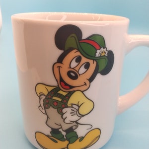 May include: White ceramic mug featuring a cartoon image of Mickey Mouse wearing a green hat and a yellow and green waistcoat. The mug has the text "DISNEY" on the bottom.