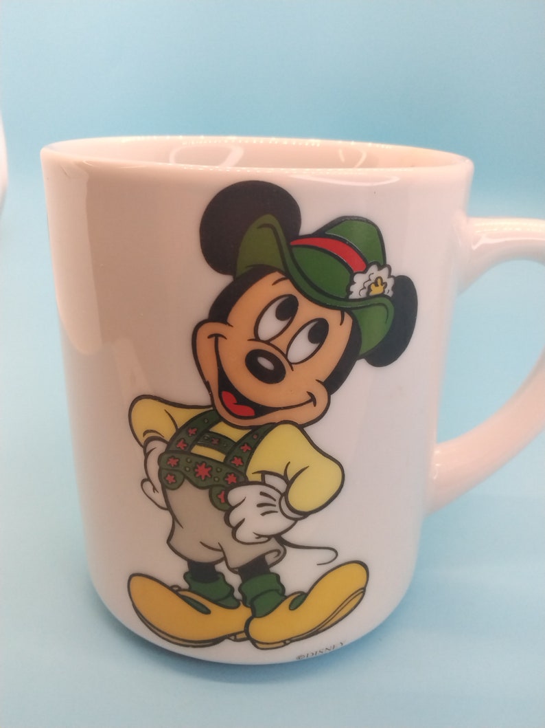 May include: White ceramic mug featuring a cartoon image of Mickey Mouse wearing a green hat and a yellow and green waistcoat. The mug has the text "DISNEY" on the bottom.