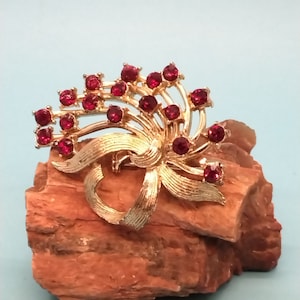 May include: A gold tone brooch with a red rhinestone design. The brooch features a bow shape with a cluster of red rhinestones in the center.