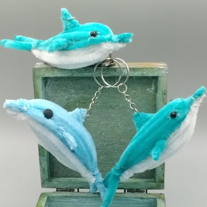 Peut inclure: Trois porte-clés dauphins en peluche bleus et blancs sont présentés. Les dauphins sont attachés à un anneau et une chaîne en métal argenté. Les porte-clés sont posés sur une boîte en bois verte patinée.
