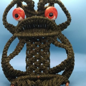May include: A green macrame frog wall hanging with a bamboo ring and red and white ceramic eyes.