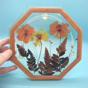 May include: A wooden framed octagonal glass coaster with pressed dried flowers and ferns. The flowers are orange, yellow, and brown. The ferns are brown.