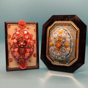 May include: Two framed decorative eggs, one with a pink and red design and the other with a blue and yellow design. Both eggs are decorated with beads and jewels.