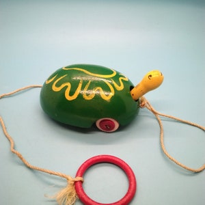 May include: A green wooden toy turtle with yellow painted lines on its shell. The turtle has a yellow head and a red wheel. The turtle is attached to a string with a red ring.