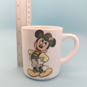 May include: White ceramic mug featuring a cartoon image of Mickey Mouse wearing a green hat and traditional German clothing. The mug has a handle and the text "DISNEY" on the bottom.