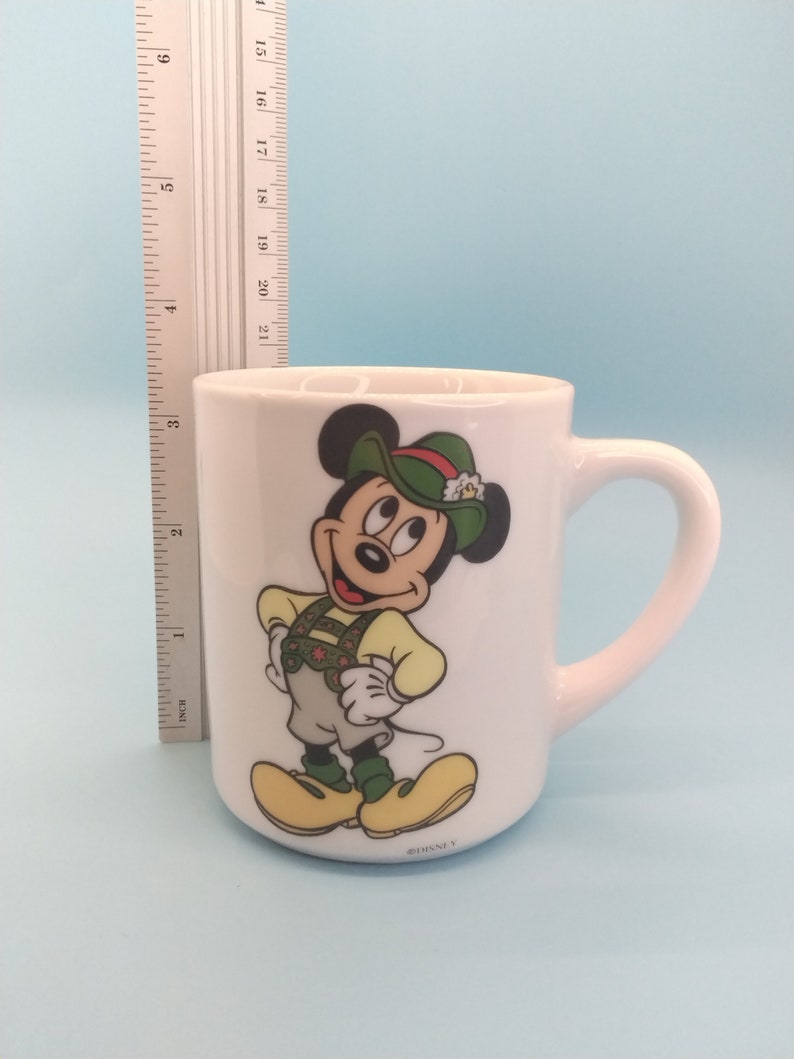 May include: White ceramic mug featuring a cartoon image of Mickey Mouse wearing a green hat and traditional German clothing. The mug has a handle and the text "DISNEY" on the bottom.