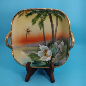 May include: A square porcelain serving dish with gold trim and handles. The dish features a hand-painted scene of palm trees, flowers, and a sunset.