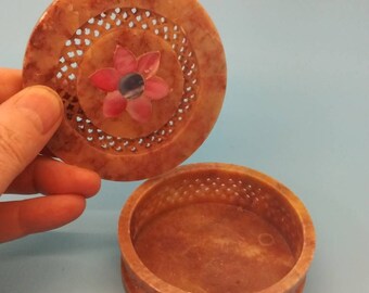 May include: A round, carved, brown stone trinket box with a floral design on the lid. The lid has a carved, openwork design and a pink and blue flower inlay.