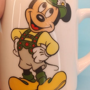 May include: White ceramic mug featuring a cartoon image of Mickey Mouse wearing a green hat and a yellow and red waistcoat. The mug has the text "&copy;DISNEY" on the bottom.