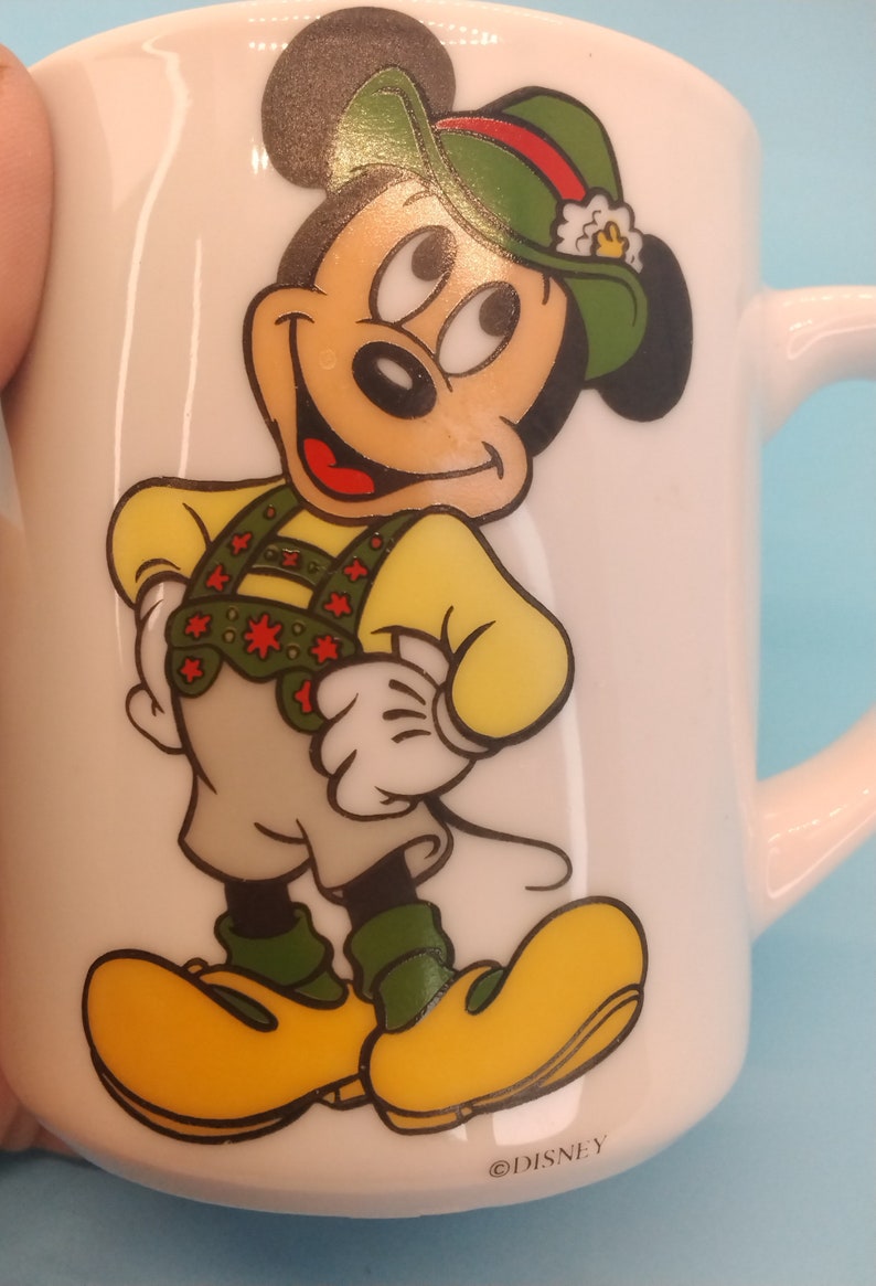 May include: White ceramic mug featuring a cartoon image of Mickey Mouse wearing a green hat and a yellow and red waistcoat. The mug has the text "&copy;DISNEY" on the bottom.