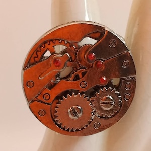 May include: A silver ring with a circular design featuring a detailed clockwork mechanism with red jewels. The ring is on a white background.