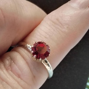 May include: A silver ring with a large, round, faceted, deep red gemstone. The ring's band has a textured pattern, and the gemstone is set in a pronged setting. The ring is shown on a finger.