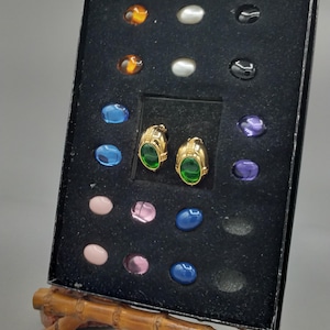 May include: A display of colourful gemstones and a pair of gold-toned earrings with green centre stones. The earrings are displayed in a black velvet-lined box, supported by a bamboo-style stand.