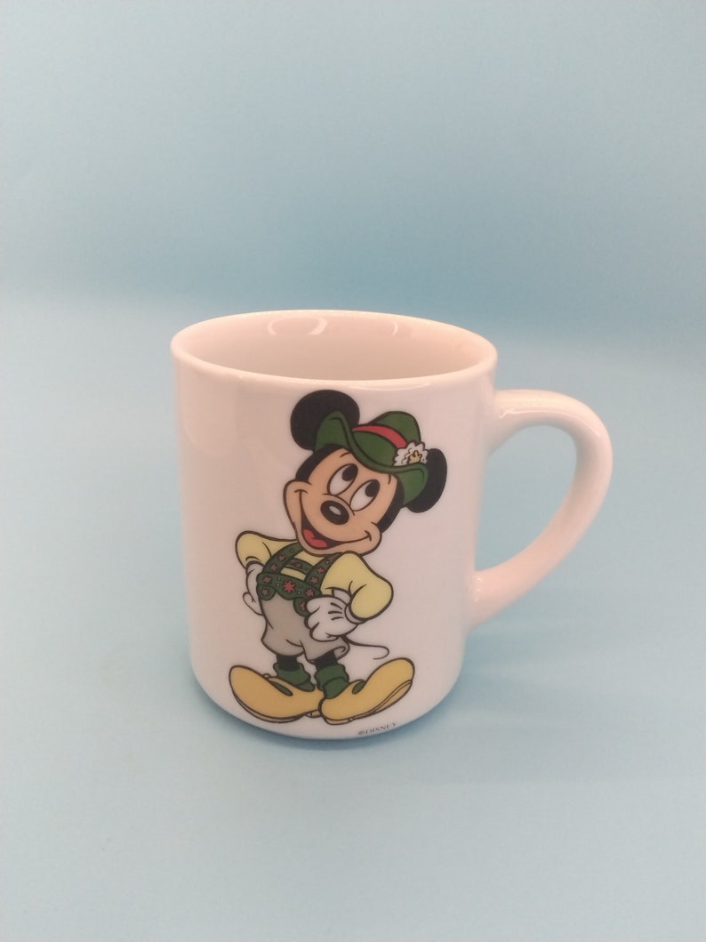 May include: White ceramic mug featuring a cartoon image of Mickey Mouse wearing a green hat and traditional German clothing. The mug has a handle and the text "&copy;Disney" on the bottom.