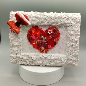May include: A decorative framed heart made of red buttons, beads, and embellishments. The heart is set against a glittery background within a white floral-textured frame. A red glitter bow adorns the frame's corner. A unique piece of art.