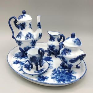 May include: A miniature porcelain tea set with a blue floral pattern. The set includes a teapot, creamer, sugar bowl, cup, saucer, and an oval serving tray. The pieces are white with blue floral designs and blue handles and accents.