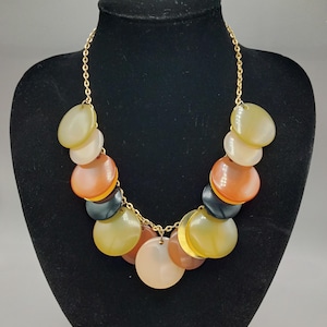 May include: A statement necklace with a gold chain and a cascade of colorful, circular pendants. The pendants are in shades of yellow, orange, black, and beige. The necklace is displayed on a black mannequin.