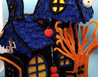 May include: A Halloween-themed craft project featuring two blue and black houses with orange pipe cleaner trees and pumpkins. The houses have windows and doors and are decorated with black pipe cleaners.