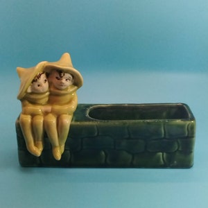 May include: A green ceramic planter shaped like a brick wall with two yellow ceramic elves sitting on top. The elves are wearing pointed hats and have brown hair.