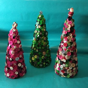 May include: Three Christmas trees made of buttons. The trees are decorated with various colours and patterns of buttons. The trees are on a turquoise background.