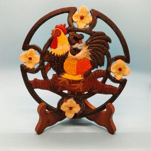 May include: A decorative rooster-themed cast iron piece, featuring a colorful rooster design with yellow, red, and brown hues. The rooster is set within a circular frame adorned with cream-colored floral accents. It is displayed on a wooden stand.