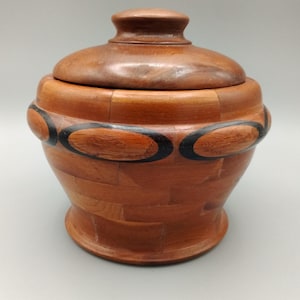 May include: A wooden container with a lid. The container is made of light brown wood with a basket weave pattern. It has a dark brown band with black oval accents. The lid has a rounded handle.