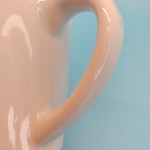 May include: A white ceramic mug with a smooth, rounded handle. The mug is glossy and has a simple design.