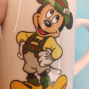 May include: White ceramic mug featuring a cartoon image of Mickey Mouse in a green hat and traditional German attire. The mug has the text "@DISNEY" printed on the base.