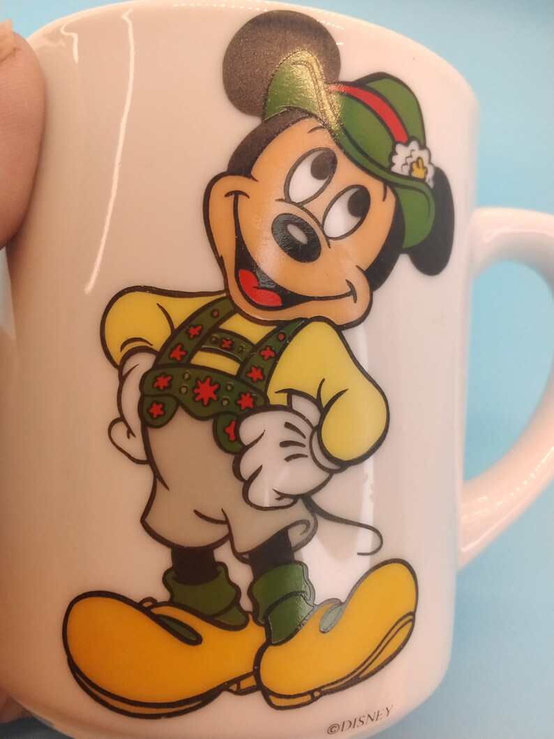 May include: White ceramic mug featuring a cartoon image of Mickey Mouse in a green hat and traditional German attire. The mug has the text "@DISNEY" printed on the base.