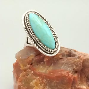 May include: A silver ring featuring an oval, light blue turquoise stone. The stone is set in a detailed bezel with a beaded border. The ring is displayed on a reddish-brown rock.