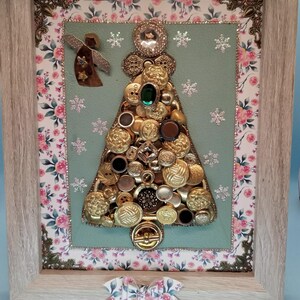 May include: A framed artwork featuring a Christmas tree made of buttons in gold, brown, and black with a green gem at the top. The frame has a floral pattern and a bow.