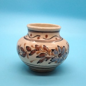 Peut inclure: Un petit vase en céramique blanche avec des motifs floraux marrons et bleus. Le vase a un corps arrondi et une ouverture étroite.