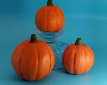 May include: Three orange pumpkins of varying sizes on a clear glass pedestal. The pumpkins are all smooth and have a green stem.