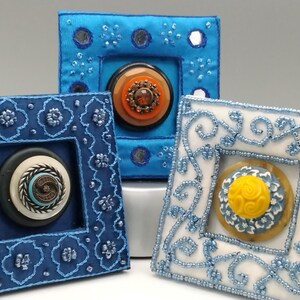 May include: Three decorative square frames with ornate button centerpieces. The frames are in shades of blue and white, with intricate beadwork and embroidery. The buttons feature various colors and designs, including orange, yellow, and black.