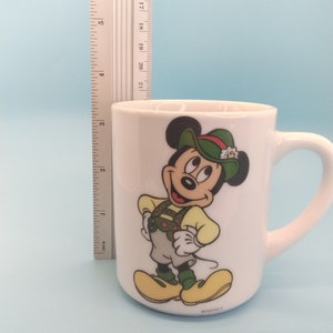 May include: White ceramic mug featuring a cartoon image of Mickey Mouse in a green hat and traditional German attire. The mug has a handle and the text "&copy;DISNEY" on the base.