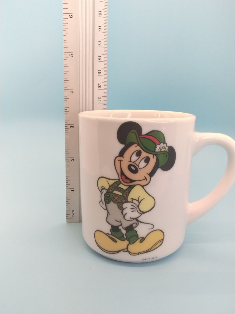 May include: White ceramic mug featuring a cartoon image of Mickey Mouse in a green hat and traditional German attire. The mug has a handle and the text "&copy;DISNEY" on the base.