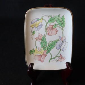 May include: A white ceramic trinket tray with a gold rim features a floral design of pink, purple, and yellow flowers with green leaves.