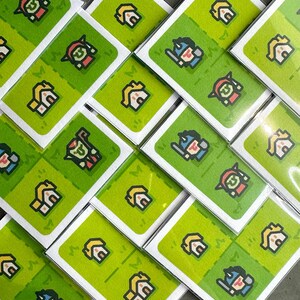 May include: A collection of white cards with green backgrounds featuring pixelated illustrations of houses, warriors, and other characters. The cards are stacked in a diagonal pattern.
