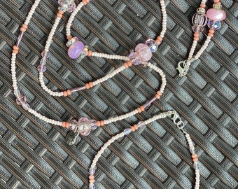Puede incluir: Un delicado conjunto de collar y pulsera con cuentas blancas y melocotón, intercaladas con cristales iridiscentes y cuentas rosas decorativas. El collar tiene un diseño largo con un cierre plateado. La pulsera es una pieza más pequeña a juego.