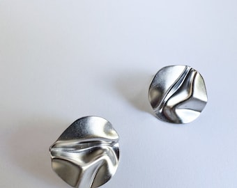 Könnte beinhalten: Ein Paar silberne Ohrringe mit zerknittertem, strukturiertem Design. Die Ohrringe sind rund und haben eine metallische Oberfläche.