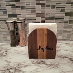 May include: A wooden napkin holder with a dark brown and light brown striped design, holding a stack of white napkins. The word "Napkins" is etched on the front. Two glass salt and pepper grinders with stainless steel tops are in the background.