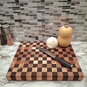 Può includere: Un tagliere in legno a scacchiera con un coltello in acciaio inossidabile, una cipolla e una zucca butternut. La tavola è composta da quadrati di legno scuro e chiaro alternati. Il coltello ha un manico nero. La zucca è marrone chiaro.