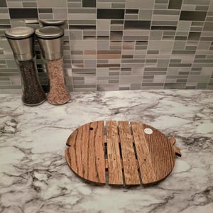 May include: A wooden fish-shaped cutting board with a natural wood grain finish. The board is made of several wooden slats and has a small hole near the head. Two glass spice grinders are in the background.
