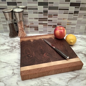 Può includere: Un tagliere quadrato in legno di testa, con un centro marrone scuro e bordi marrone chiaro. Un coltello in acciaio inossidabile è appoggiato sul tagliere, accanto a una mela rossa e un limone giallo. Due macinini per sale e pepe in vetro sono sullo sfondo.